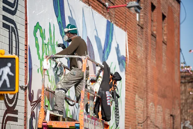 An Open Air Gallery painter working from a lift platform, mask on, part way through a wall